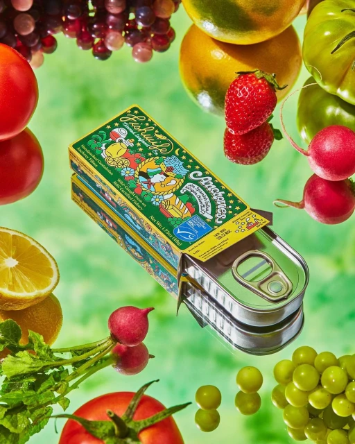 Artistic food still life featuring an open tin of anchovies, tomatoes, lemons, grapes, and radishes on a glossy green surface.