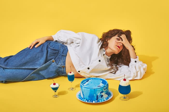 A woman with curly hair lies on a yellow background with blue cake and dessert glasses. She wears jeans and a white button-down shirt.