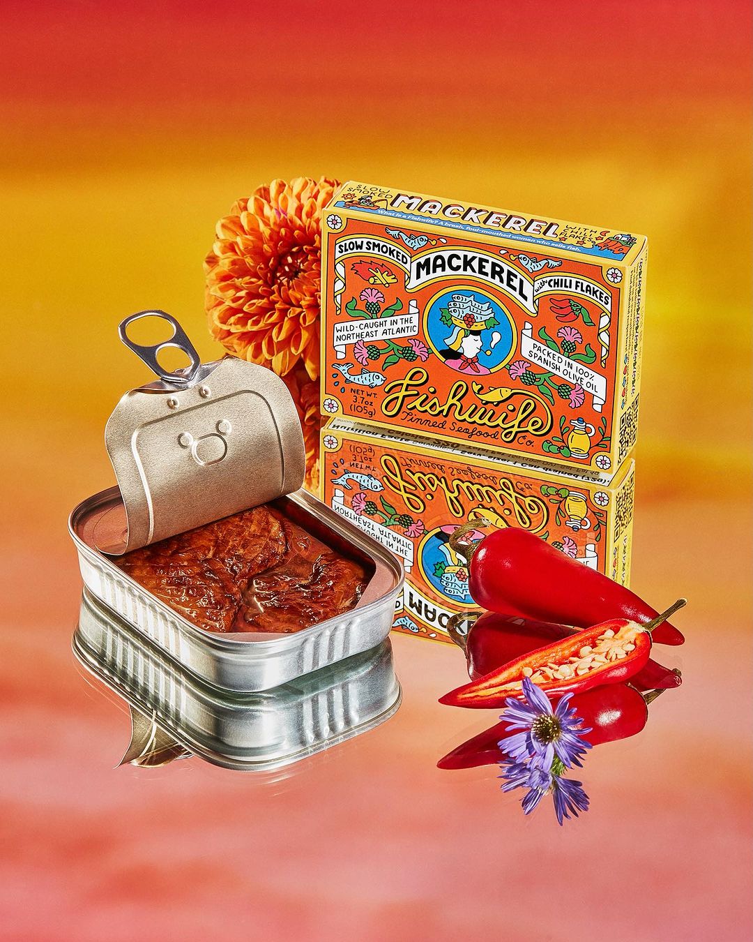 Canned mackerel product photography on a reflective surface with flowers and peppers against an orange and yellow background.