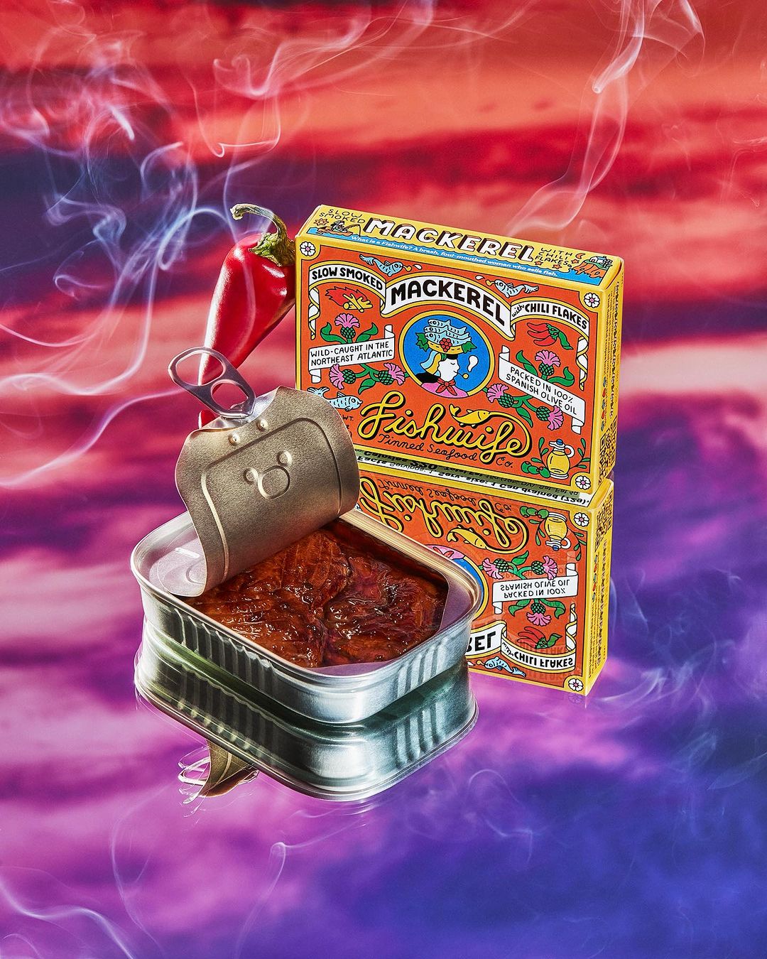 Canned mackerel fillets in a tin, next to stacked product boxes and a fresh red chili, against a vibrant abstract background.