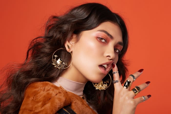 Close-up portrait of a woman wearing large ornate hoop earrings, multiple rings, and red eyeshadow against an orange background
