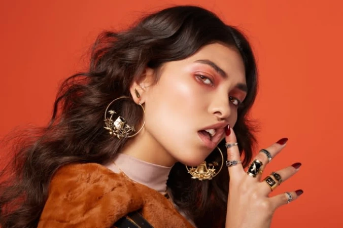 Close-up portrait of a woman wearing large ornate hoop earrings, multiple rings, and red eyeshadow against an orange background