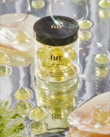 Fur Bath Drops jar sits amongst shell fragments and bubbles. The jar is clear with a black top and filled with yellow bath drops.
