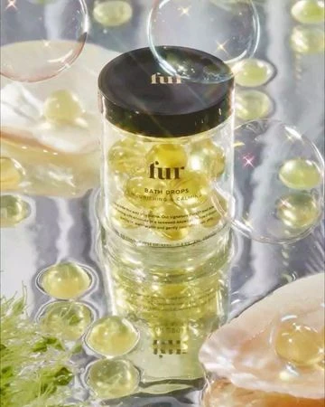 Fur Bath Drops jar sits amongst shell fragments and bubbles. The jar is clear with a black top and filled with yellow bath drops.