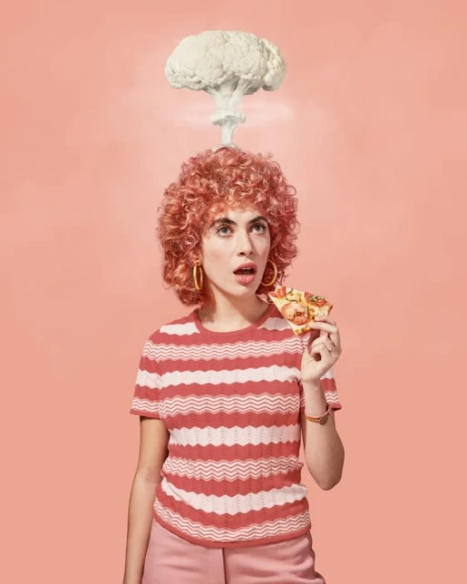 Young woman with curly red hair eats pizza slice under an atomic cloud of cauliflower against a peach background in a studio shot.