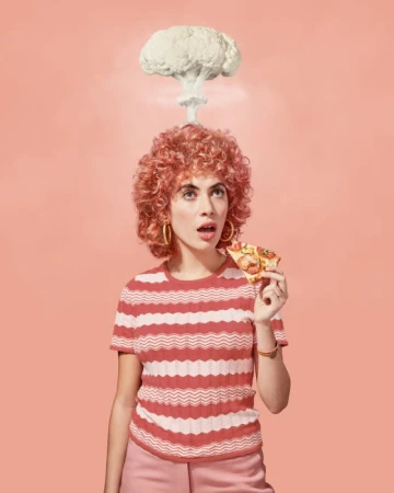 Young woman with curly red hair eats pizza slice under an atomic cloud of cauliflower against a peach background in a studio shot.