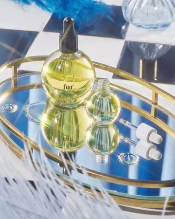 Cosmetic product still life: two glass bottles of oil and dropper caps on a gold tray, with white feathers in the foreground.