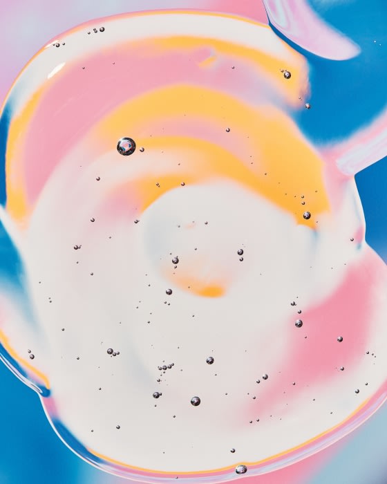 Close-up shot of a translucent cosmetic gel with pink, yellow, and white swirls, and small scattered air bubbles.