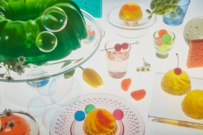 Overhead shot of vibrant green and yellow gelatin desserts on clear glass dishes, complemented by delicate flowers and bubble accents.