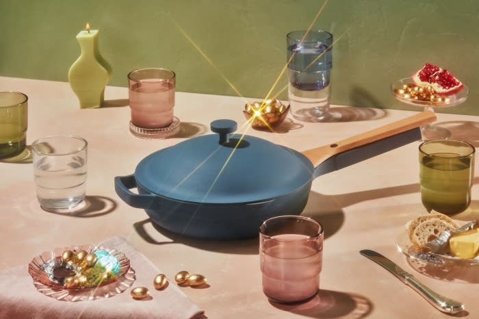 Still life featuring a blue Always Pan with a lid, surrounded by glasses, a candle, and small plates with snacks on a pink table.
