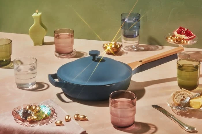 Still life featuring a blue Always Pan with a lid, surrounded by glasses, a candle, and small plates with snacks on a pink table.