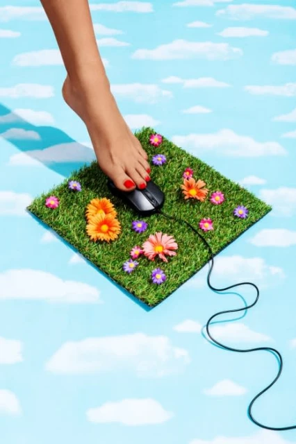 A bare foot presses down on a computer mouse, which sits on a square of artificial grass decorated with flowers, against a blue cloudy sky.