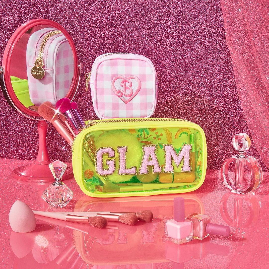 Glamorous beauty product presentation featuring makeup bags, brushes, nail polish, and perfume in a pink setting with glitter backdrop.