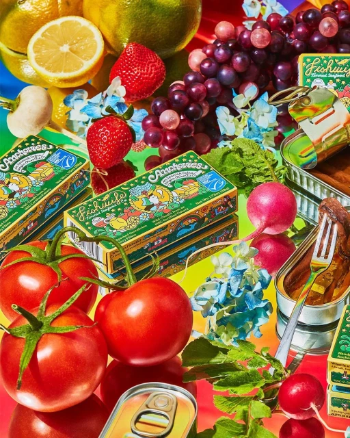 Still life arrangement with tinned fish, tomatoes, strawberries, radishes, grapes, lemons, and canned sardines, creating a colorful food composition.