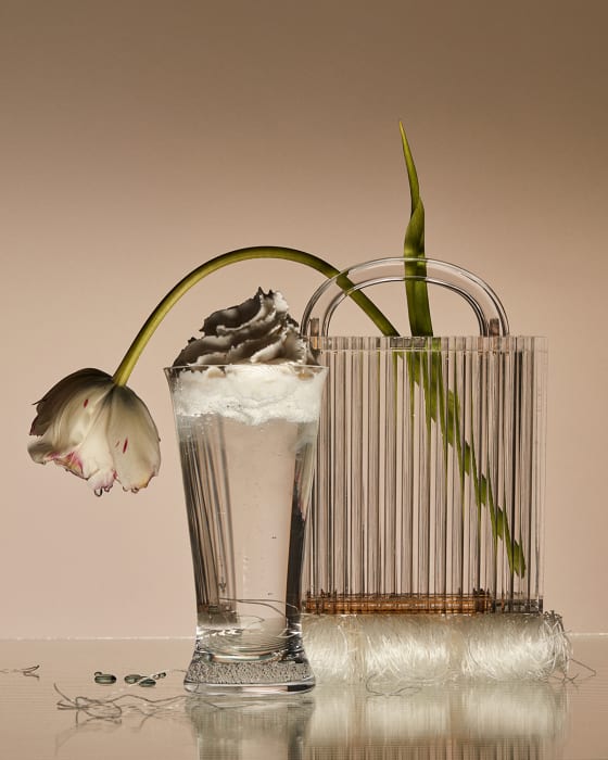 Tulip flower head hangs over an elegant glass filled with water and topped with grey foam. Clear bag behind it.