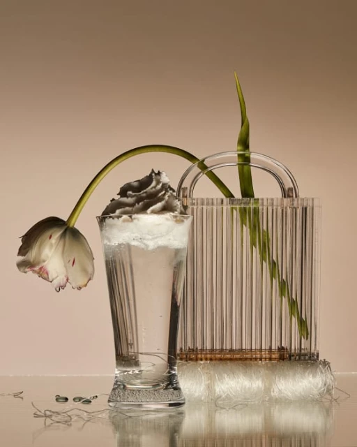 Tulip flower head hangs over an elegant glass filled with water and topped with grey foam. Clear bag behind it.