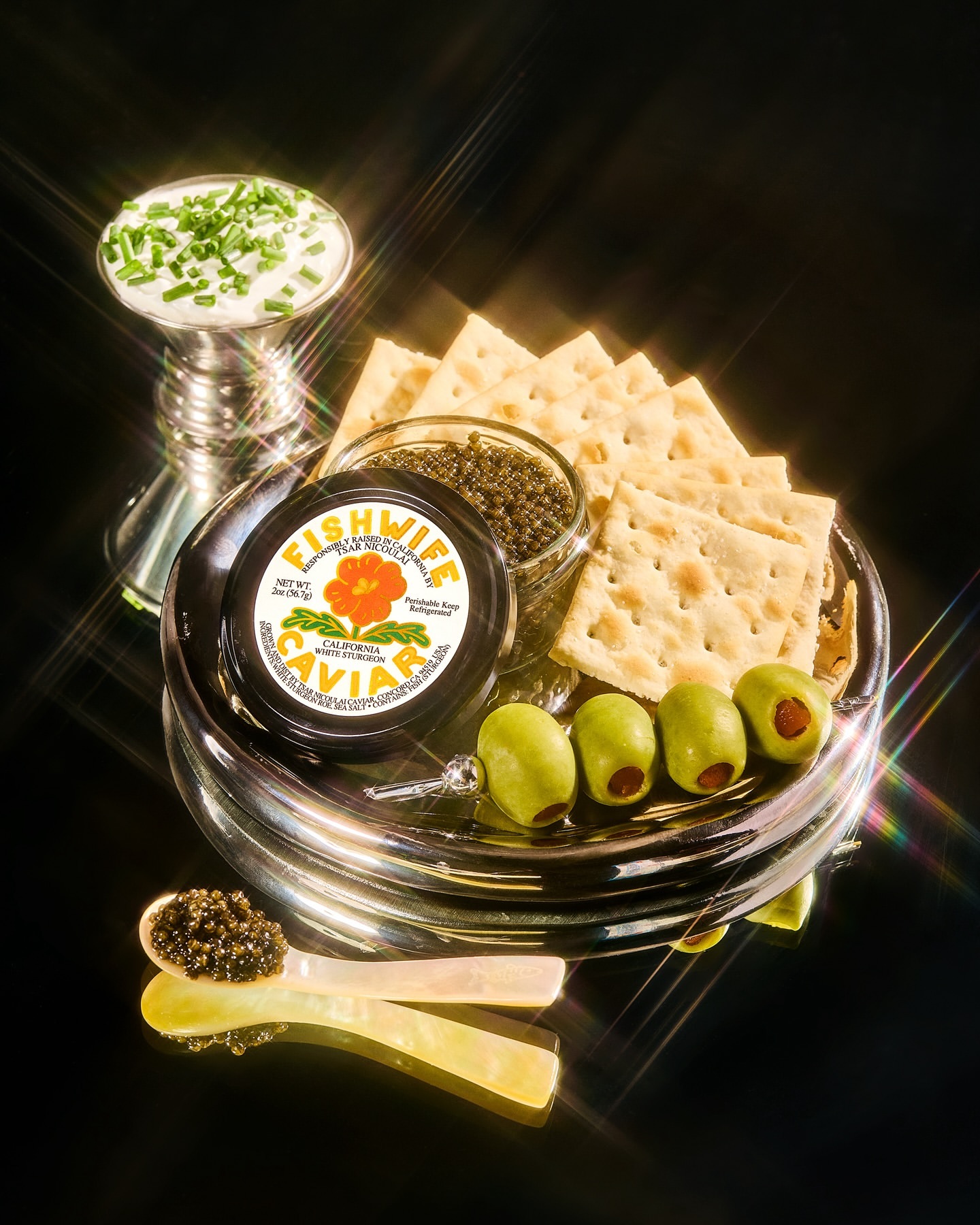 A still life presents Fishwife caviar with crackers, olives, and crème fraîche on a reflective black surface, showcasing gourmet food photography.