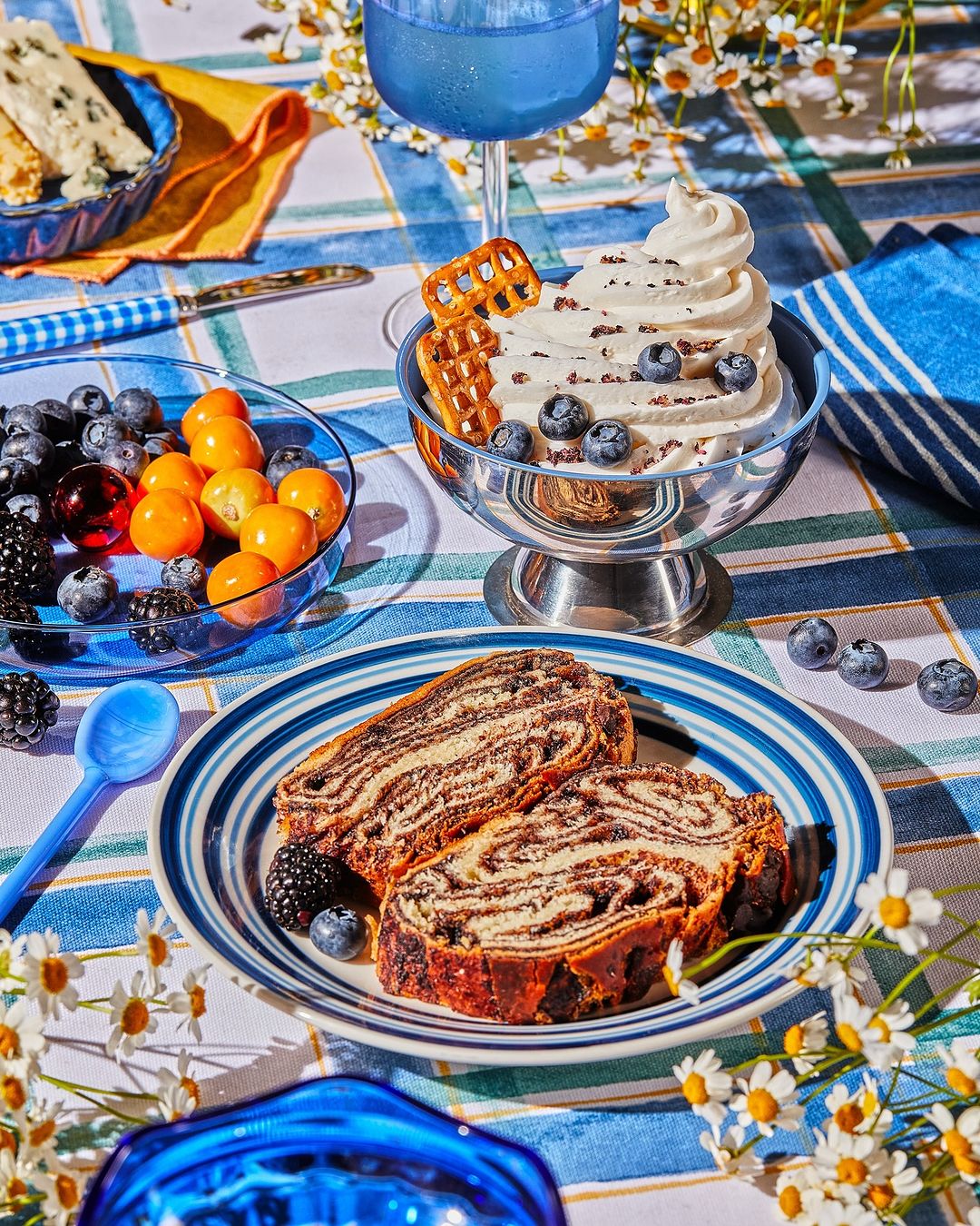 Overhead food photography of sliced babka on a plate with berries, ice cream, and a drink, set on a blue striped tablecloth.