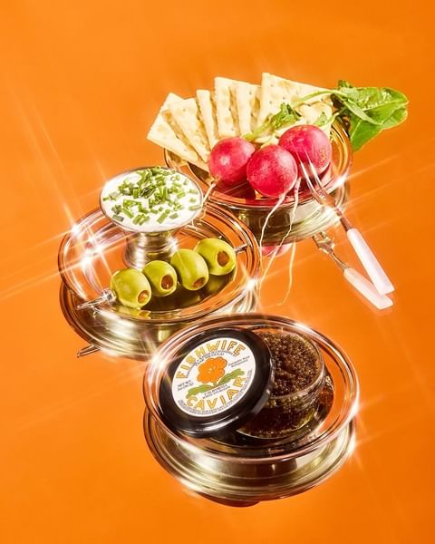 Caviar and appetizers served on silver stands and plates against an orange background, featuring radishes, crackers, olives, and chives.