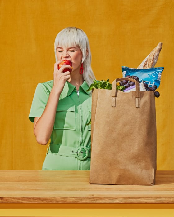 Woman with platinum blonde hair in green jumpsuit eating red apple in front of a brown grocery bag filled with various foods.