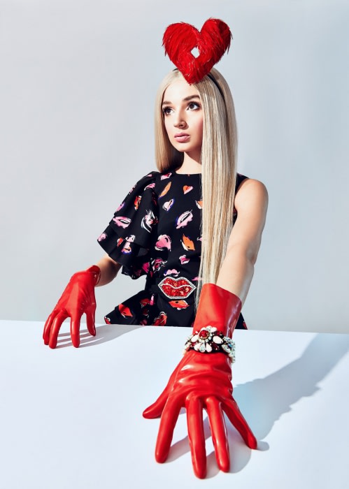 Woman with heart headpiece and red gloves posing in a studio. Blonde hair, patterned dress, bright colors against a plain background.