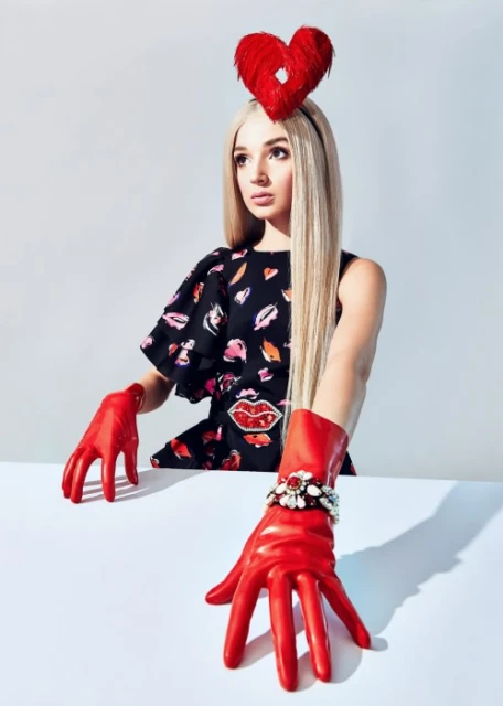 Woman with heart headpiece and red gloves posing in a studio. Blonde hair, patterned dress, bright colors against a plain background.