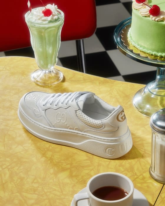 Gucci sneaker sits on a diner table with a milkshake, cake, coffee and a sugar dispenser in a retro diner setting.