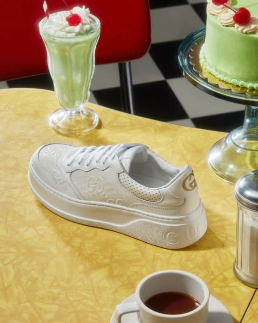 Gucci sneaker sits on a diner table with a milkshake, cake, coffee and a sugar dispenser in a retro diner setting.