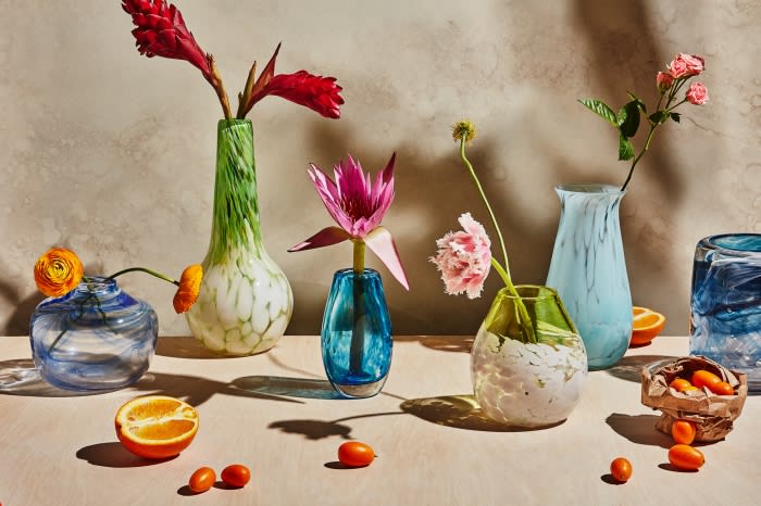 Still life with colorful glass vases holding flowers, alongside sliced oranges and kumquats, set against a neutral mottled background.