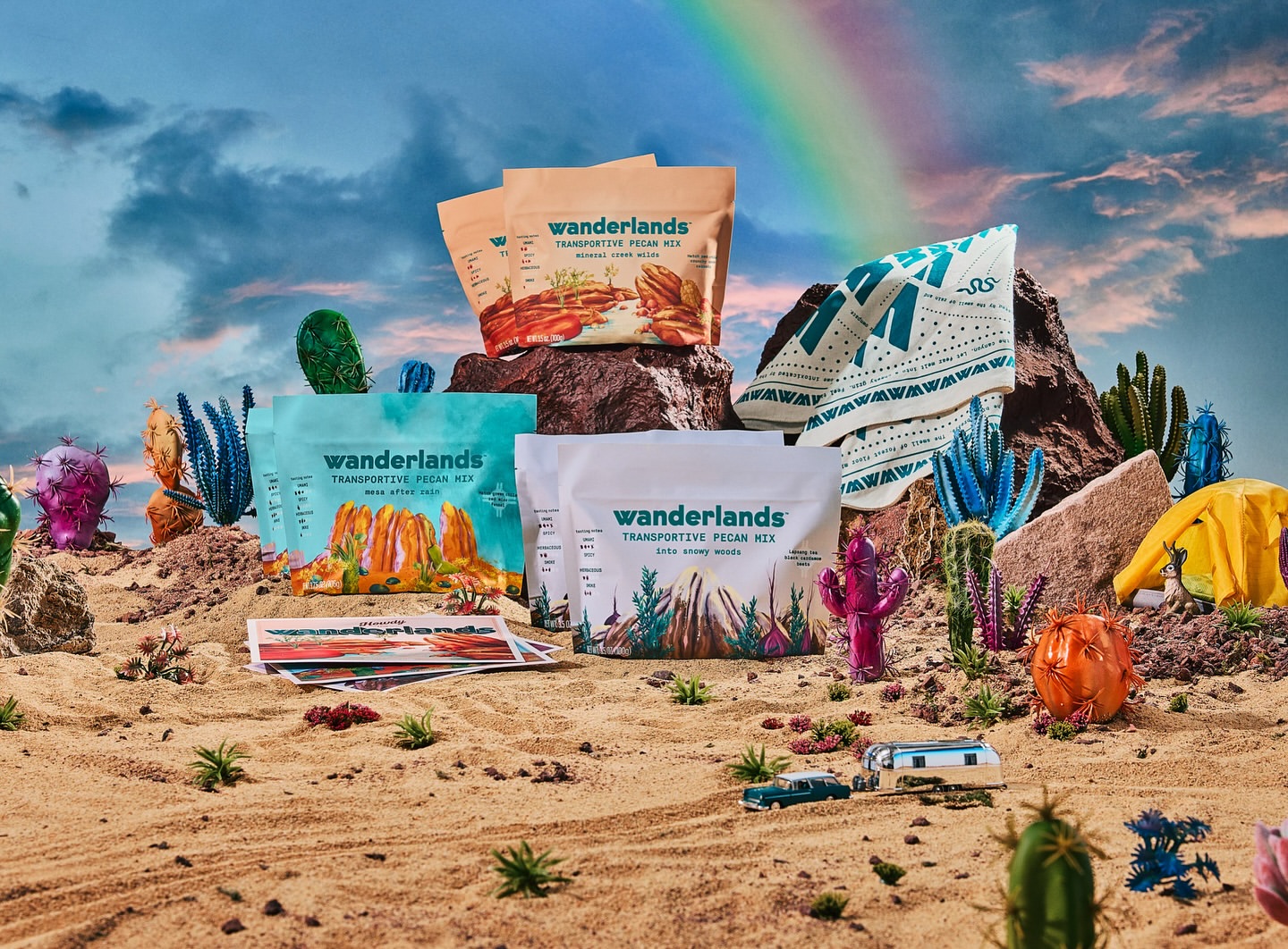 Product display of Wanderlands Transportive Pecan Mix in a desert scene with cacti, rocks, toy car, and rainbow against a cloudy sky.