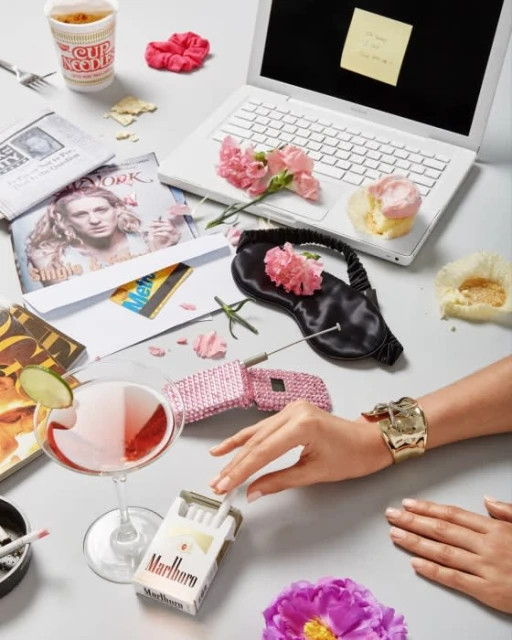 Still life on a table featuring a hand, cocktail, cigarettes, laptop, beauty products, and food items in bright, eye-level studio shot.