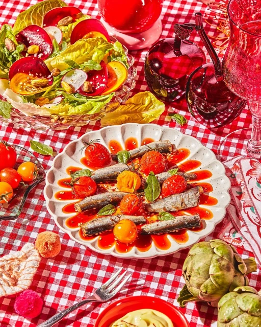 Overhead shot of sardine dish with tomato sauce, garnished with basil, salad, red drink on a red and white checkered tablecloth.