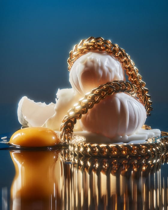 Close-up of a cracked chicken egg with yolk, egg white, and shell fragments, adorned with a chunky gold chain. Studio setting, reflective surface.