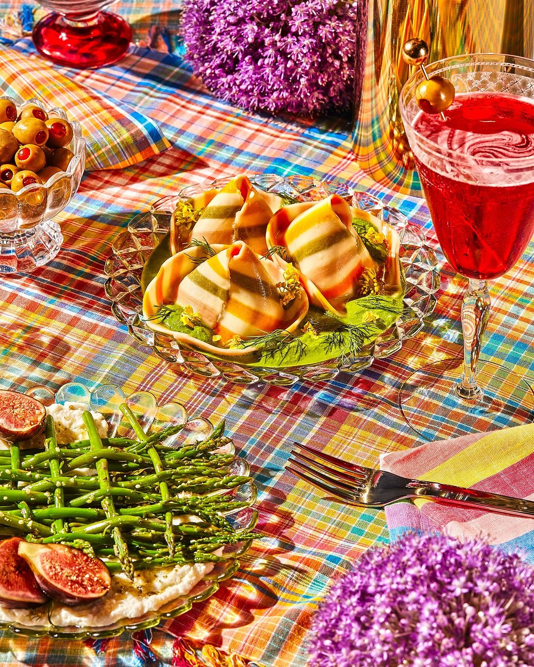 Overhead shot of a crystal table setting featuring asparagus, figs, appetizer rolls, olives, and a red cocktail, all on a colorful tablecloth.