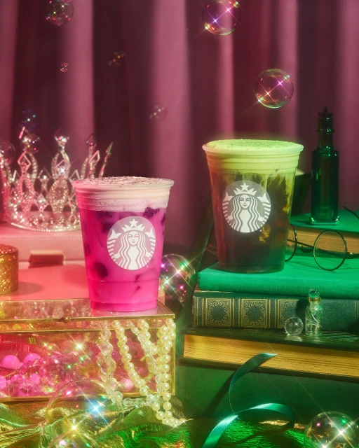Two Starbucks beverages, one pink and one green, sitting on a stack of books with fairy lights and bubbles around them in a studio setting.