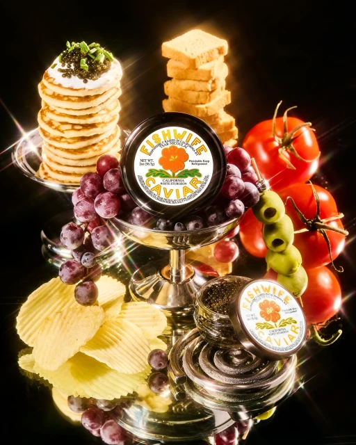 Still life featuring Fishwife brand California white sturgeon caviar with grapes, tomatoes, potato chips, pancakes, and toast on a reflective surface.