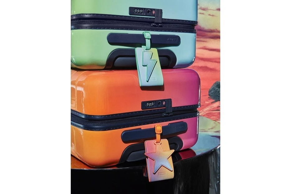 Stack of colorful hard shell suitcases with a lightning bolt and star luggage tag in a studio setting against a desert background.