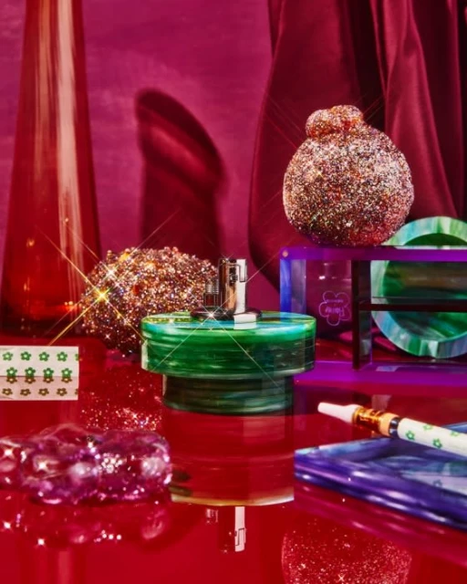 Product shot featuring a perfume bottle with a metallic top, surrounded by glittery spheres and reflective surfaces on a red backdrop.