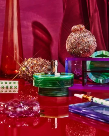 Product shot featuring a perfume bottle with a metallic top, surrounded by glittery spheres and reflective surfaces on a red backdrop.