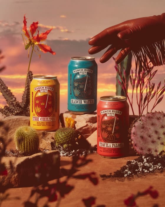 Three Ranch Rider canned cocktails, including Chilton, Ranch Water, and Tequila Paloma, rest on stones with desert plants against a sunset sky.