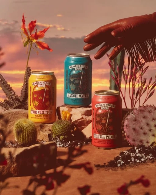 Three Ranch Rider canned cocktails, including Chilton, Ranch Water, and Tequila Paloma, rest on stones with desert plants against a sunset sky.