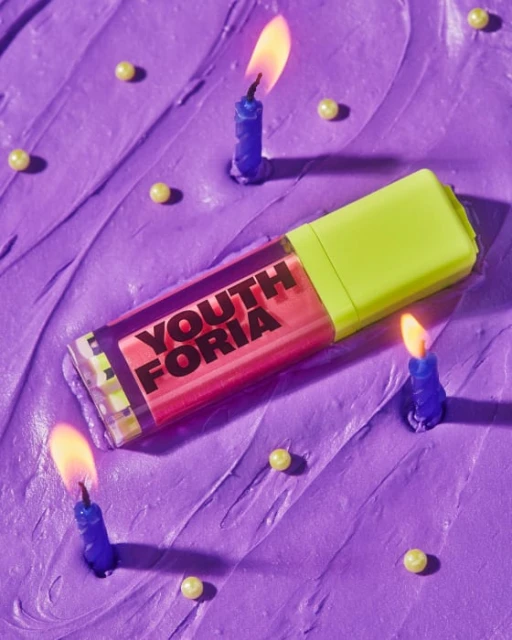 Cosmetic product with a lime green cap sits on a purple cake surface surrounded by small blue birthday candles with lit flames.