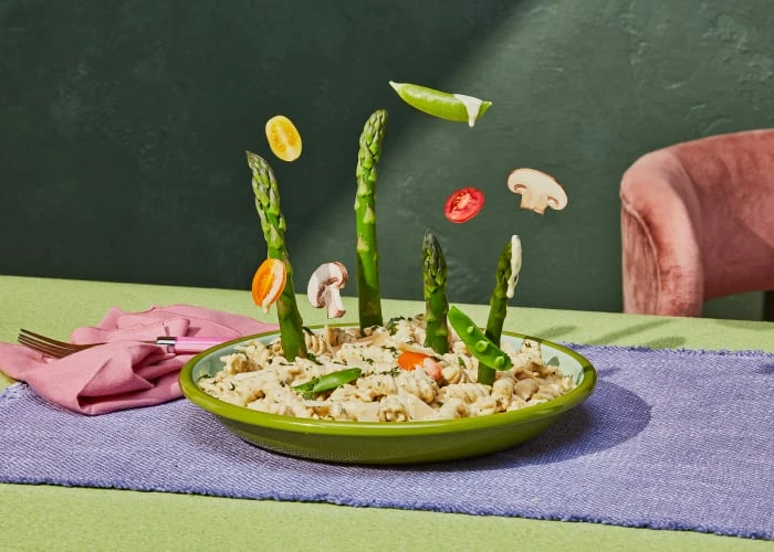 Overhead shot of green bowl of pasta with asparagus, peas, mushrooms, tomato, and squash arranged on a table with green backdrop.