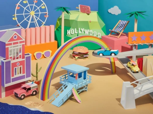 Whimsical paper craft scenery featuring a rainbow, cars, buildings, palm trees, lifeguard tower, burger, and Hollywood sign against a blue sky.