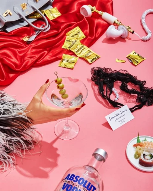 Flat lay photo of a martini glass with olives, condom packets, purse, Absolut vodka bottle, phone and sushi on a pink surface.