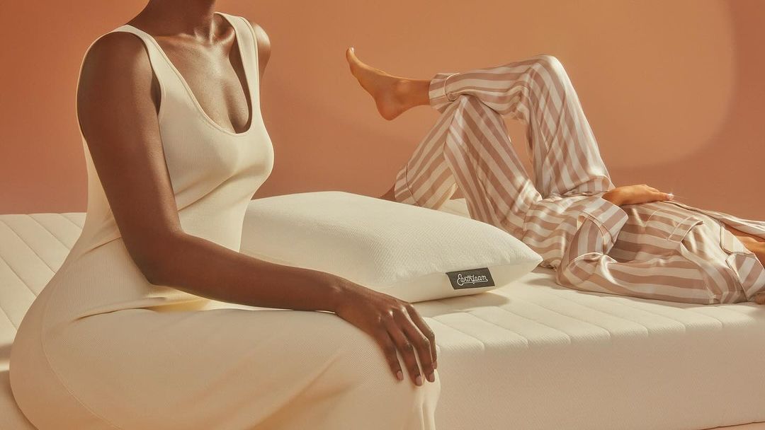 Two women, one sitting and one reclining, showcase a mattress and pillow in a warm-toned, indoor setting, emphasizing comfort.