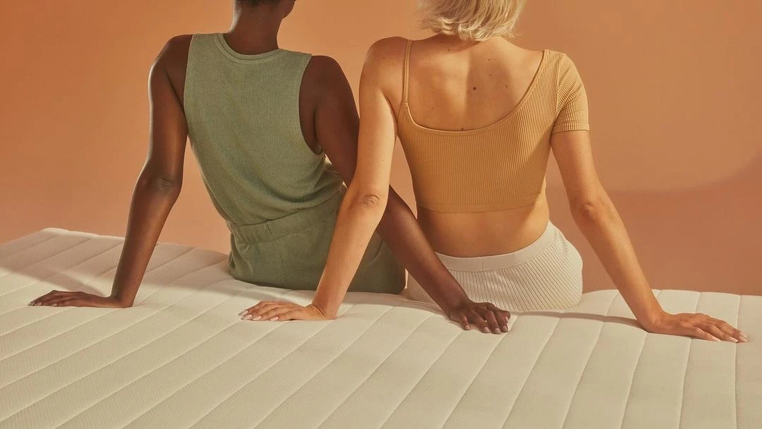 Two people are sitting on a white mattress in a studio. The two people have their backs to the camera in a back lit studio.