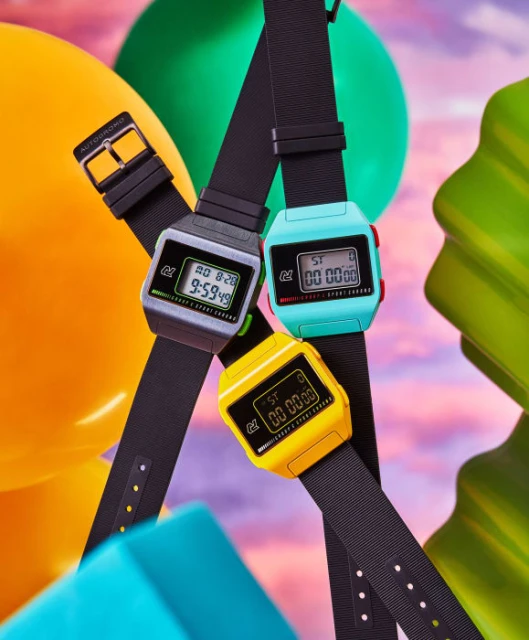 Three digital wristwatches are displayed in grey, turquoise, and yellow against a balloon and cloud-painted background, focused on the product.