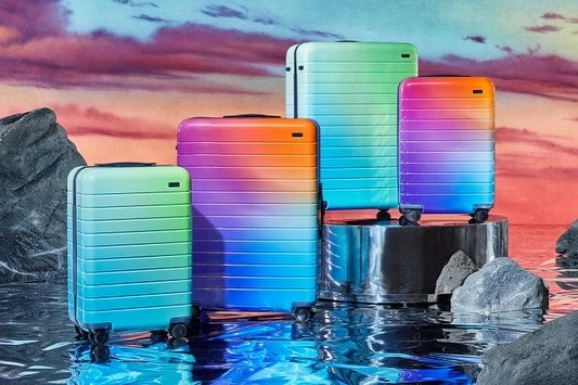 Four colorful, ridged suitcases in a row, staged in a surreal, watery setting with rock formations and a pink-blue sunset sky backdrop.