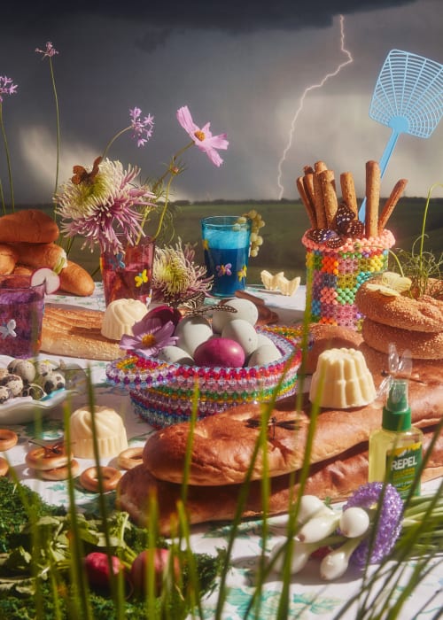 Table laden with bread, drinks, eggs, and floral arrangements under a stormy sky with visible lightning strikes in the background.